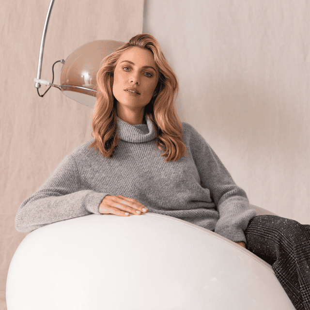 Image of a woman sat on a white puff. She's wearing a gray rolled neck jumper, and gray trousers