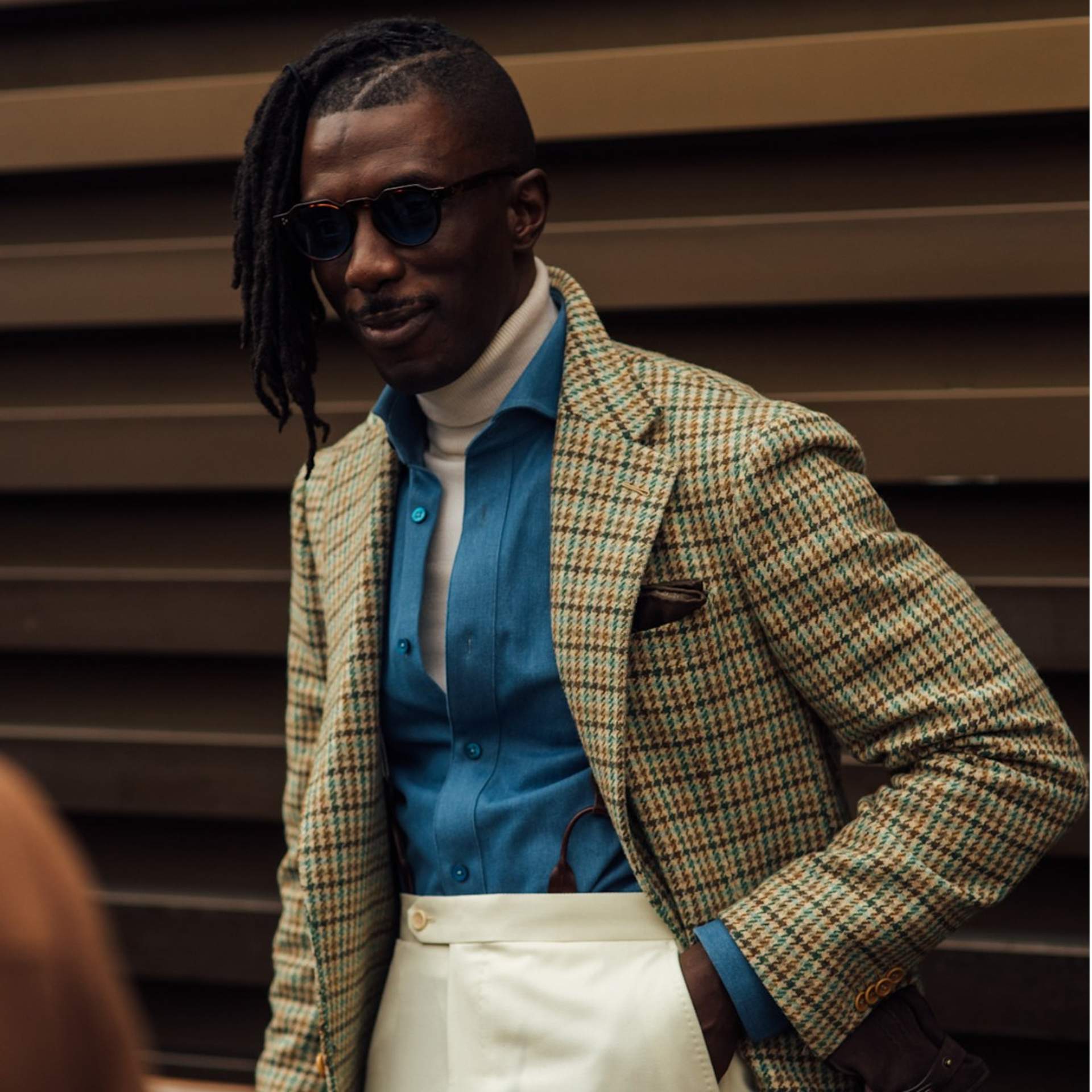 A man showcasing a stylish look with cream suit trousers, a cream knitted roll neck with a blue chambre on top then a tweed blazer as a final layering piece 