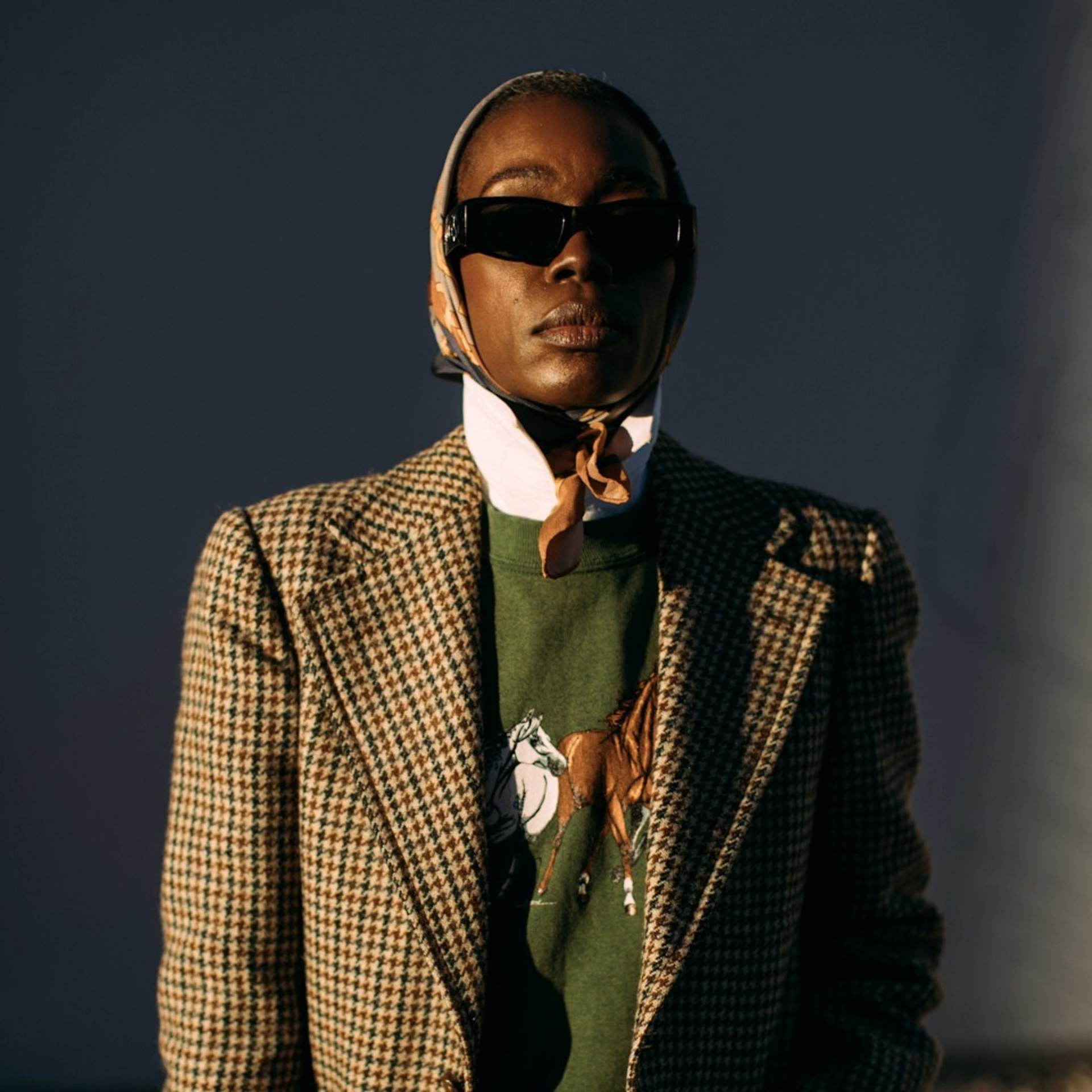 A woman showcasing a layered outfit consisting of a treed oversized blazer with a green jumper with embroidered horses then a white shirt underneath with the collar popped-up and a headscarf to finish off the look. 