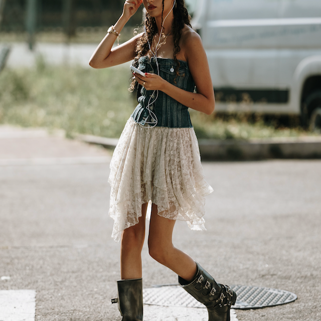 a young woman wearing a festival style outfit of a white boho lace style skirt, black biker boots and a corset style top. 