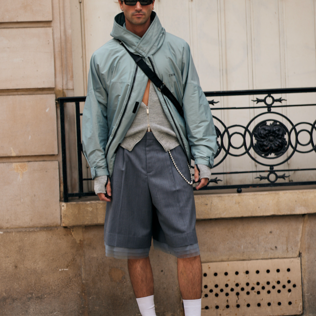 a male standing in front of a beige stone wall, wearing a bomber jacket, long denim shorts and accessories, streetwear style
