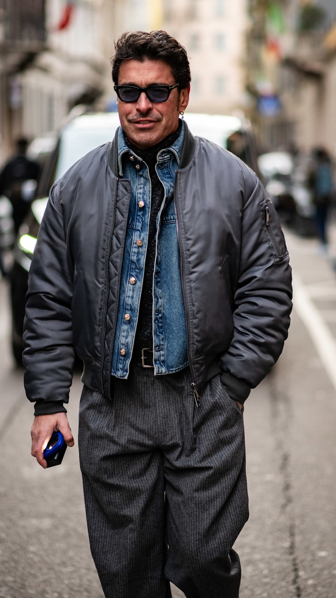 A man with an overszied grey metallic bomber jacket with a light blue wash denim jacket underneath with a roll neck base layer
