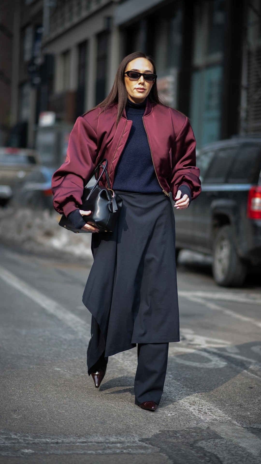 A woman walking whilst wearing a long pleated skirt, a blue knitted rollneck and an oversized maroon bomber with a metallic finish