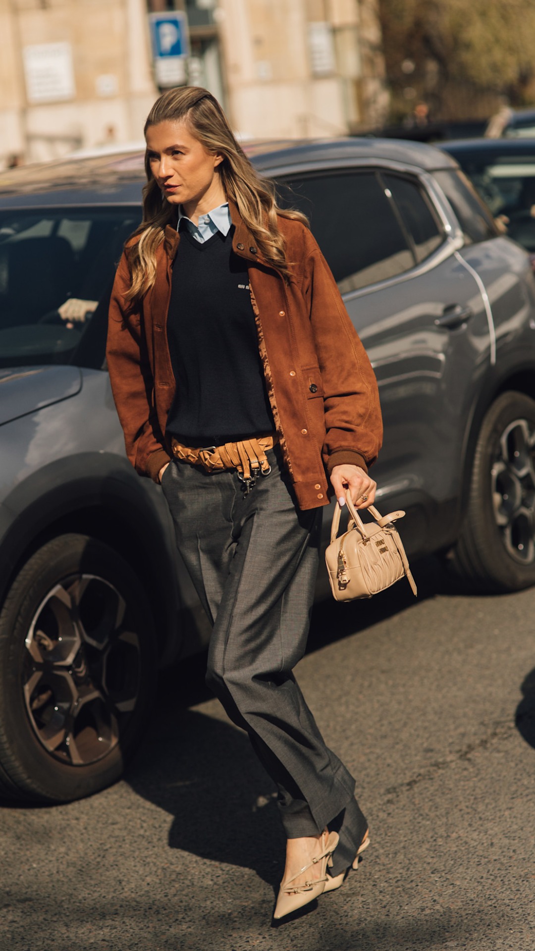 A woman walking whilst wearing a layered outfit consisting of dark baggy jeans, a light brown suede jacket with a blue knitted v-neck sweater and light blue poplin shirt layered underneath