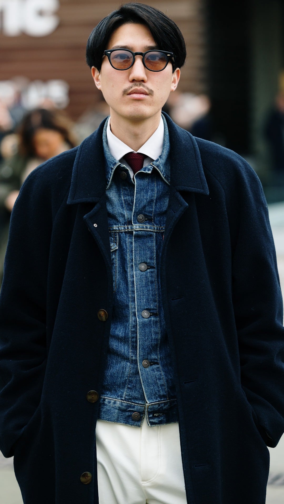 A man with several clothing layers on including a stylish black coat, a light blue wash denim jacket with a white shirt and tie underneath
